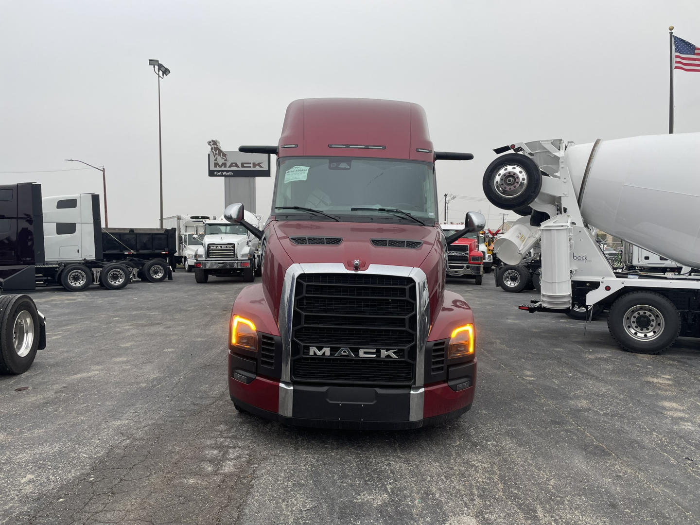 Truck front view in a showroom lot showing red Mack PR64T76H sleeper cab with large black grille and illuminated headlights.