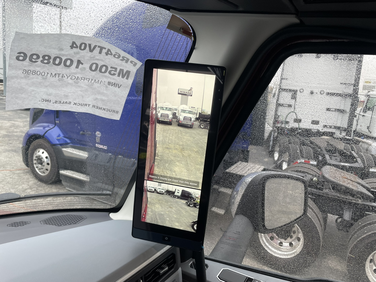 Truck cab interior showing a vertical touchscreen display mounted near the windshield with side mirrors and other trucks visible outside.