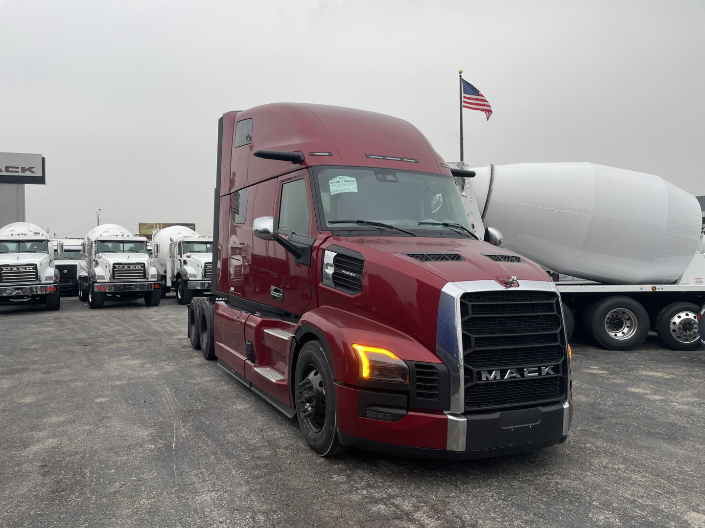 Mack PR64T76H truck in a dealership lot, maroon cab with black grille, large sleeper, front view with headlights on, adjacent white fuel tank trailer in background.