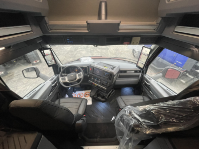 Mack PR64T76H cabin interior showing dashboard with steering wheel and center console, driver seat and passenger seat in view, plastic seat wrap on right, overhead storage, large side windows.
