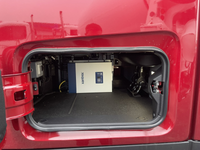 Front storage compartment of a red Mack PR64T76H truck showing a white Xantrex power inverter in the bay.