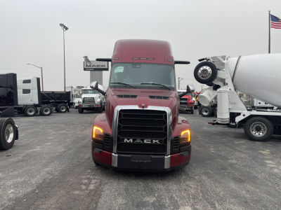 Truck front view in a showroom lot showing red Mack PR64T76H sleeper cab with large black grille and illuminated headlights.