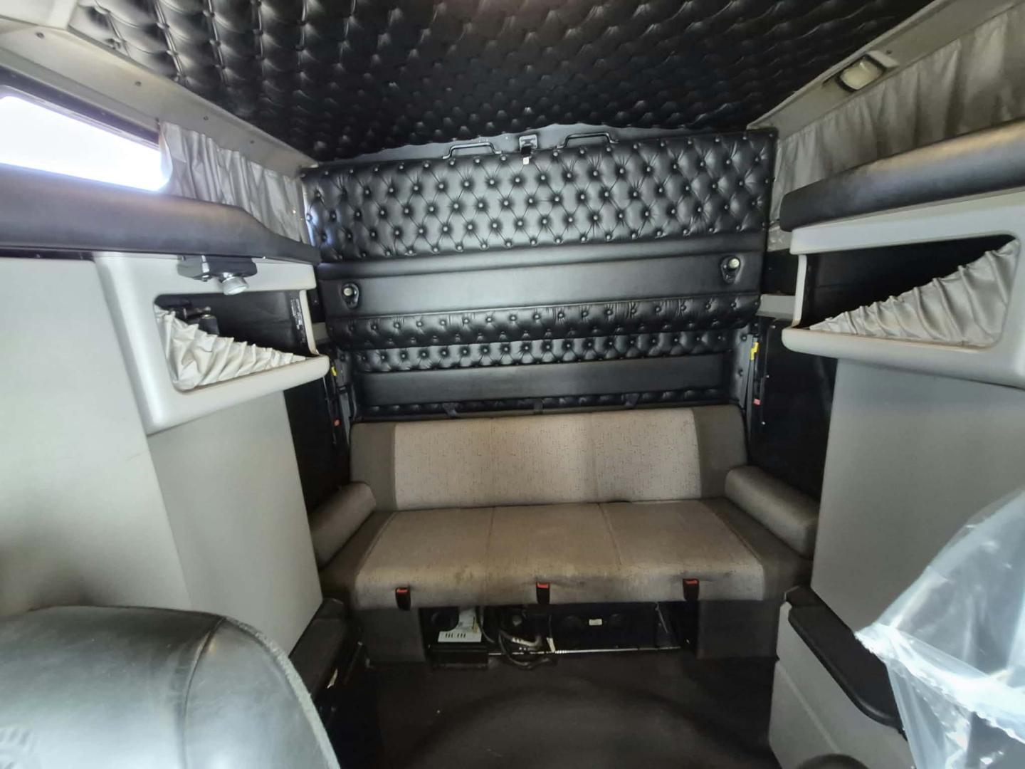Kenworth W900L truck cab interior showing quilted black padded ceiling and walls, beige bunk seat, and overhead storage shelves.