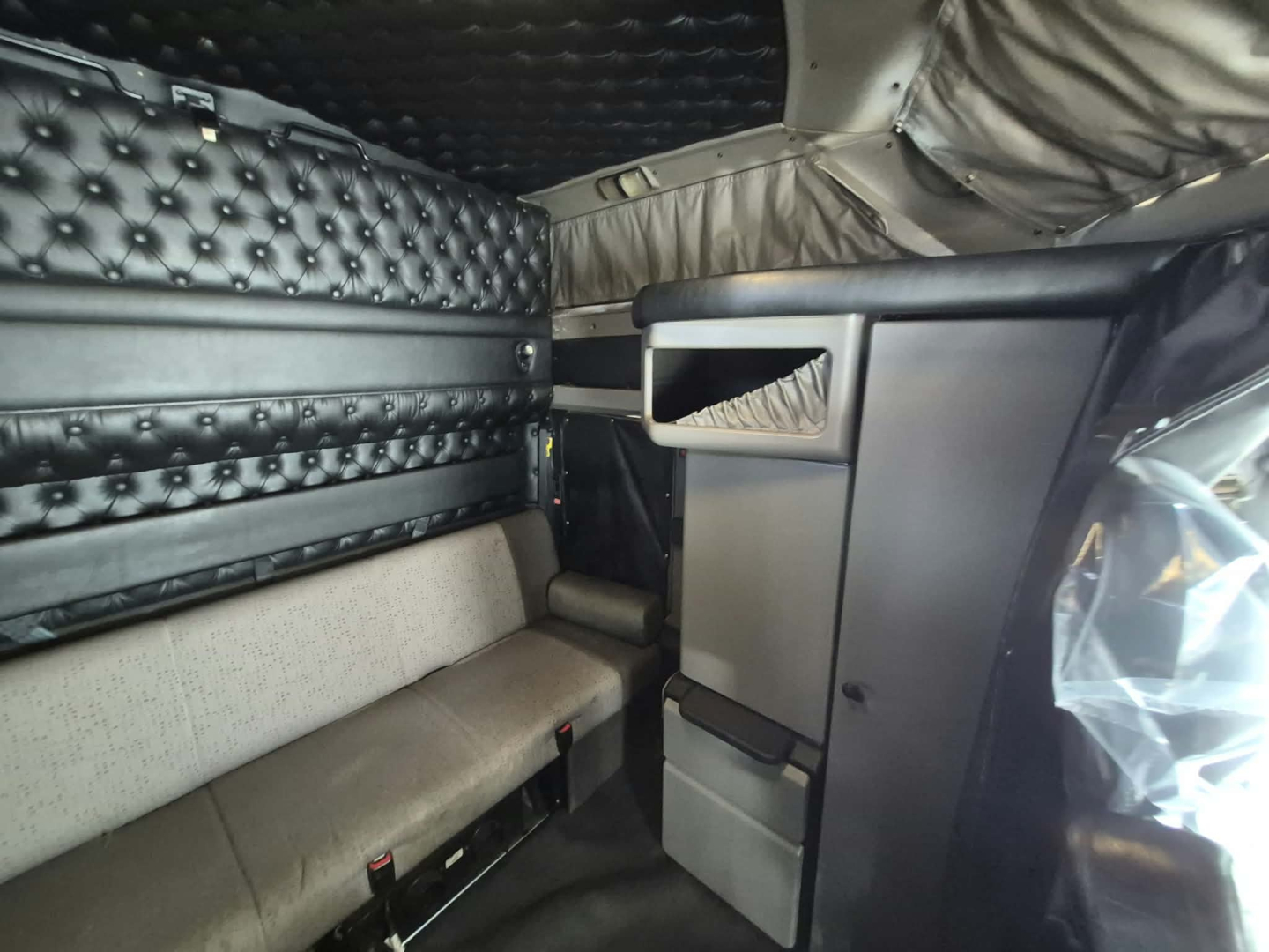 Inside Kenworth truck cab with quilted black air-spring partition wall, grey upholstered bench, storage compartment and overhead panels visible.