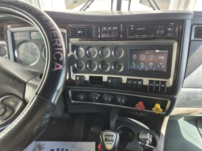 Kenworth W900L dash area showing steering wheel with 'AMERICAN' text, multiple gauges, touchscreen display, switches, and controls.