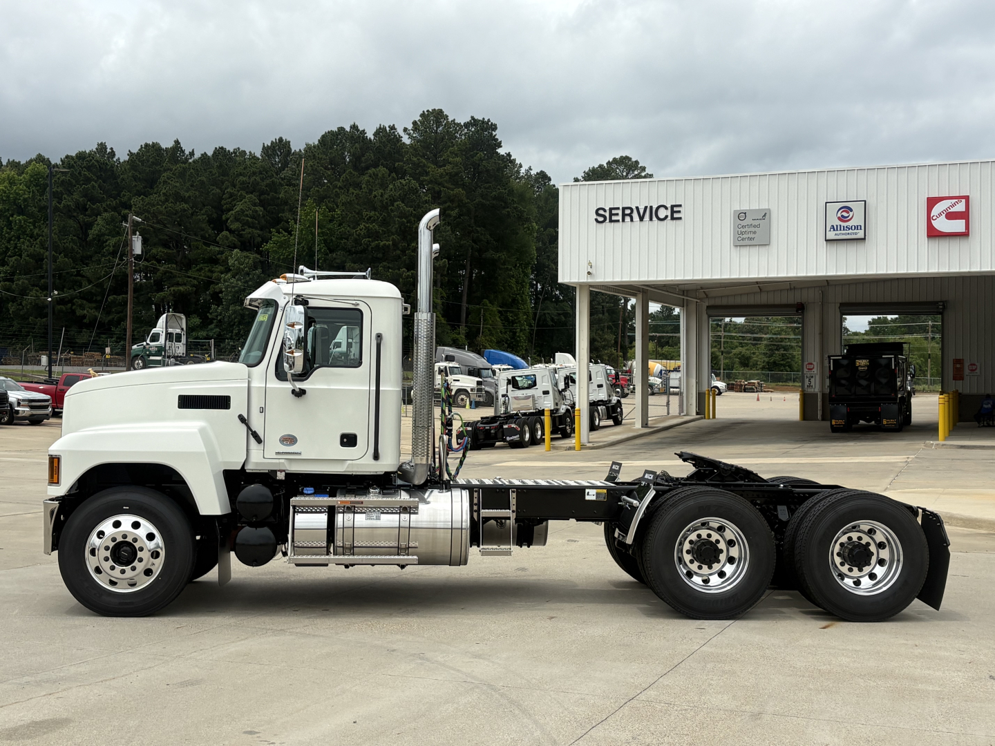 Truck cab and chassis of a Mack PI64T in a dealership lot, side view showing air intakes, fuel tanks, and rear wheel axles, service bay in background.