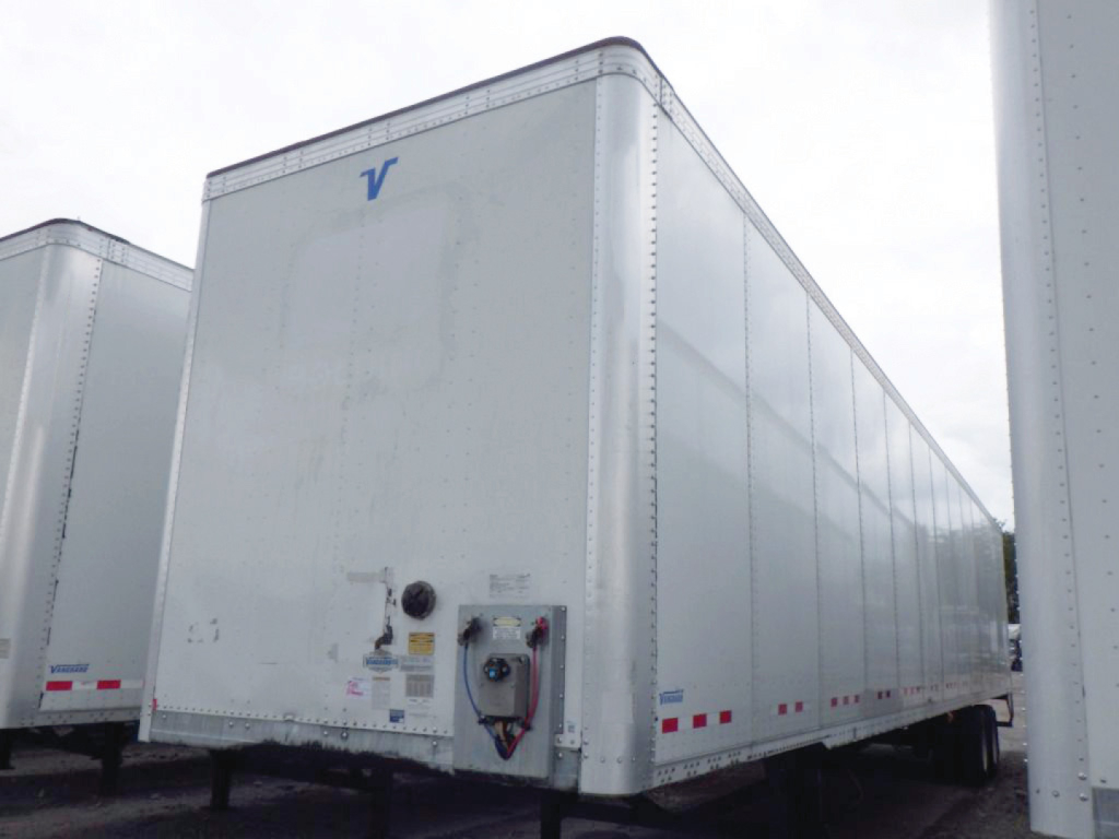 Trailer exterior showing a white Vanguard VXP box trailer with a blue 'V' logo on the front corner, side access door, and reflective tape along lower edge.