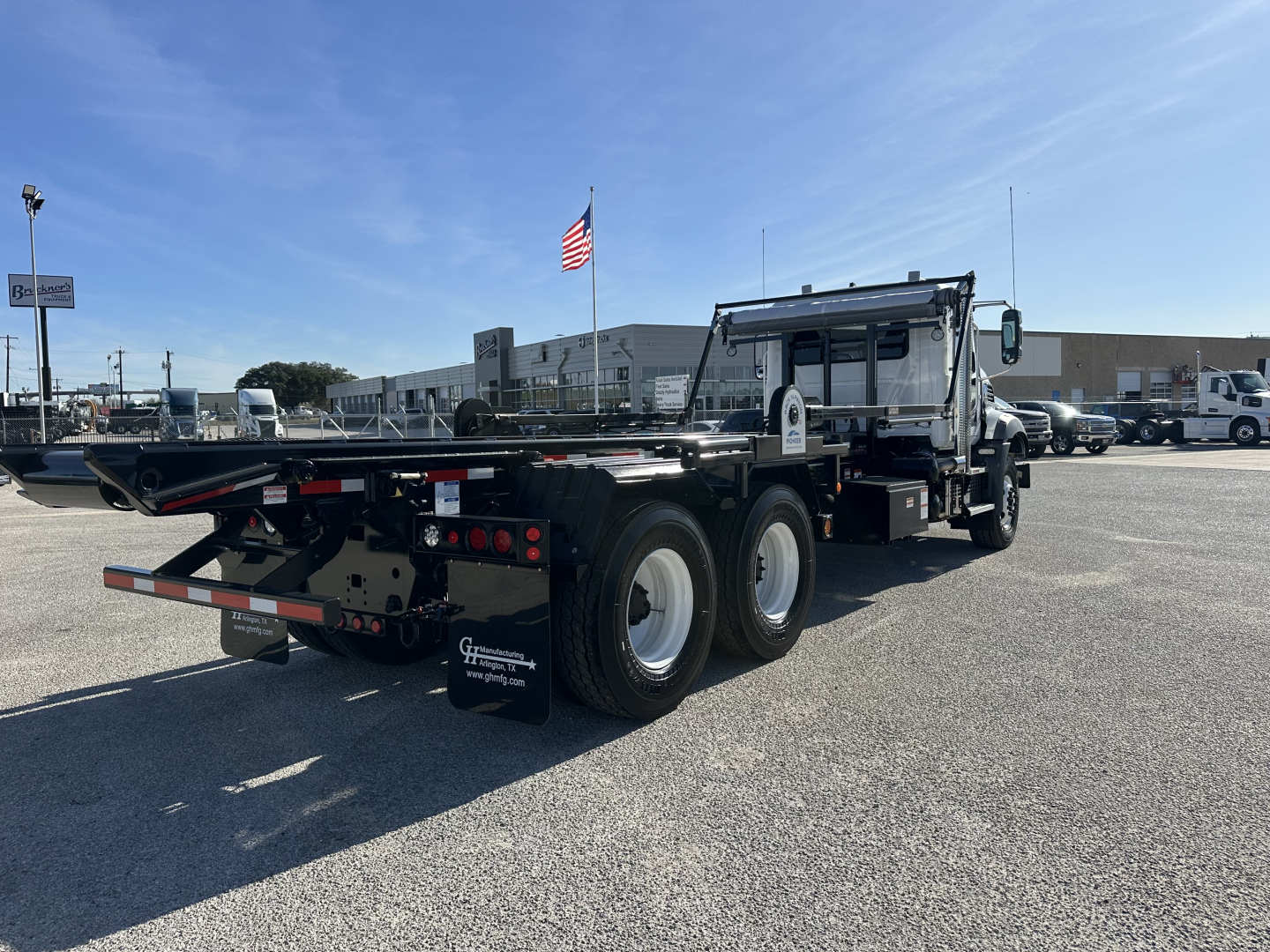 Mack GR64B rolloff truck in a parking lot, three rear axles, black flatbed, cab with chrome details, American flag in background.