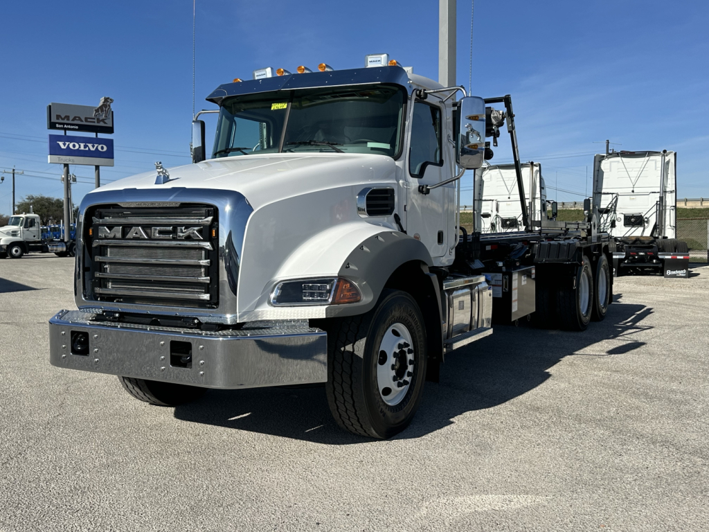 Mack GR64B roll-off truck in white cab with chrome bumper and flatbed, front grille reading MACK, DVIN visible in background.