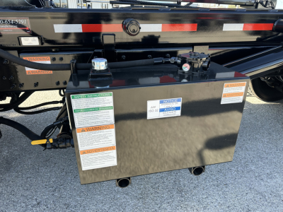 Truck component showing a fuel or hydraulic tank with warning labels and caps, mounted behind the cab of a 2026 Mack GR64B roll-off truck.