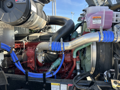 Engine bay of a 2026 Mack GR64B roll-off truck showing red engine block, blue silicone hoses, aluminum intercooler piping, pink coolant reservoir, and various labels.