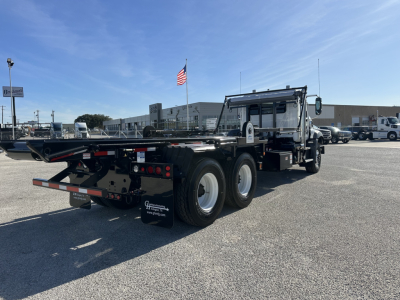 Mack GR64B rolloff truck in a parking lot, three rear axles, black flatbed, cab with chrome details, American flag in background.