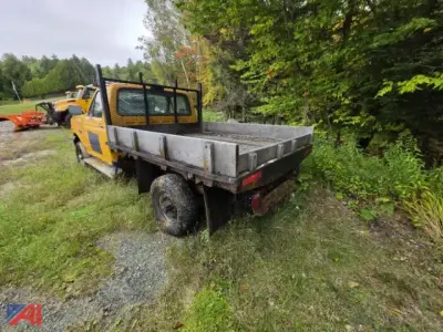 Flatbed tow truck with yellow cab and metal flatbed, rear view showing towing bed and off-road setting.