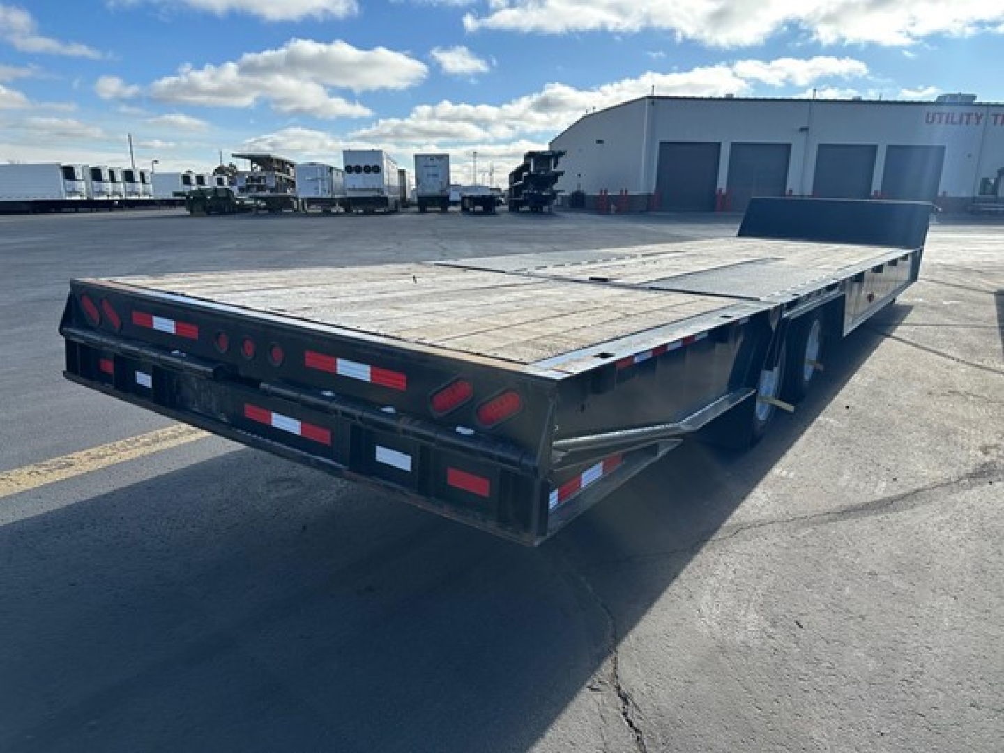 Trailer bed showing flatbed trailer with black steel frame and wooden deck, rear view with taillights and reflective tape, utility yard in background.