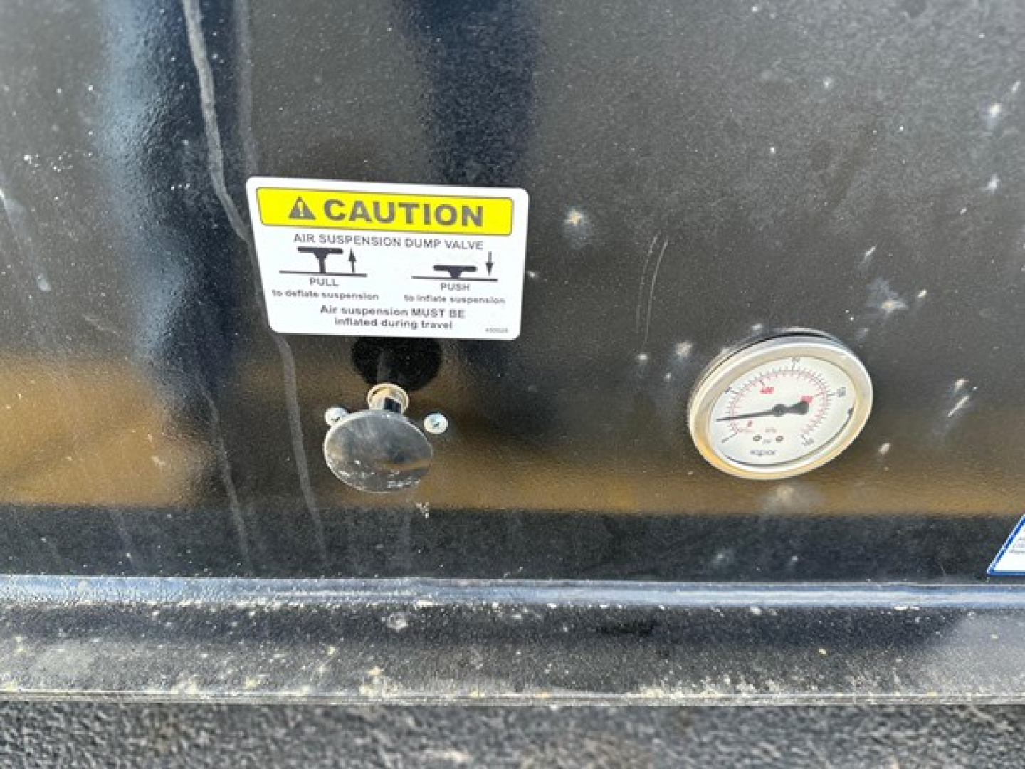 Caution label about air suspension dump valve and gauge on trailer front panel; small round gauge; black surface with D-shaped valve knob.