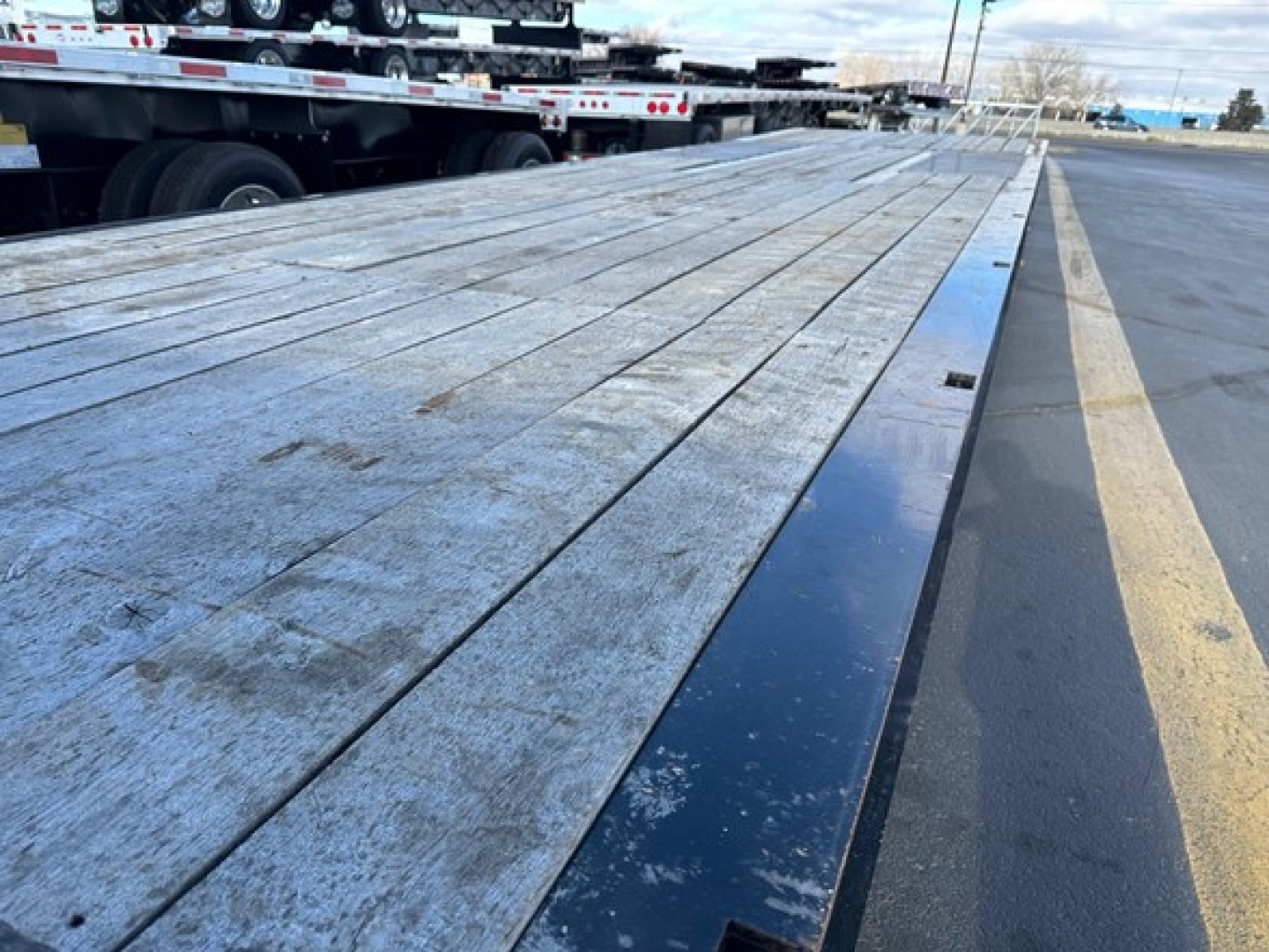 Flatbed trailer deck with weathered wooden planks and a blue steel edge rail; part of a truck trailer visible on left, road and parking lot in background.