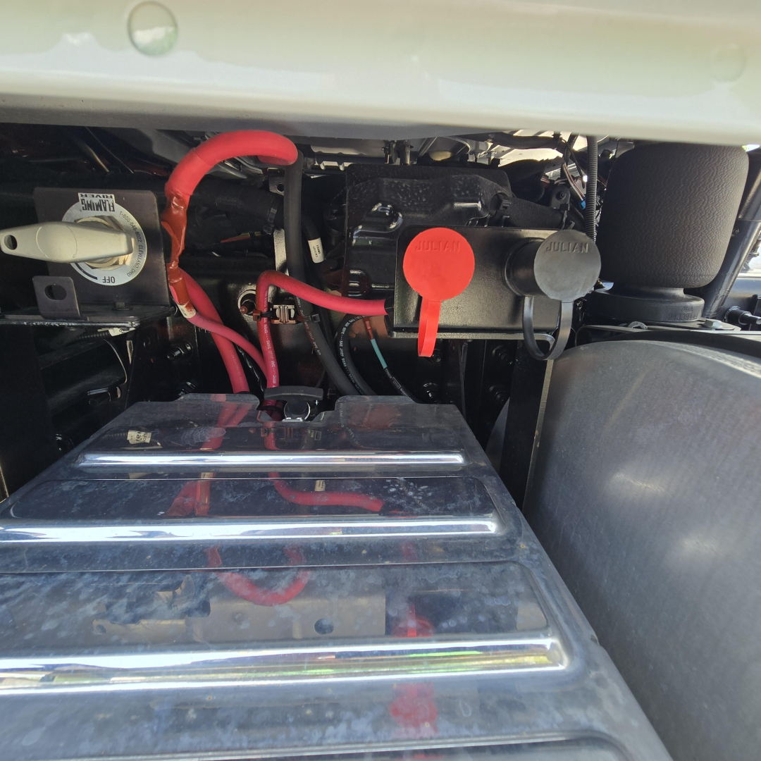 Mack GR64B engine bay showing battery cover and red/leaded hoses with dipstick and coolant reservoir cap visible.