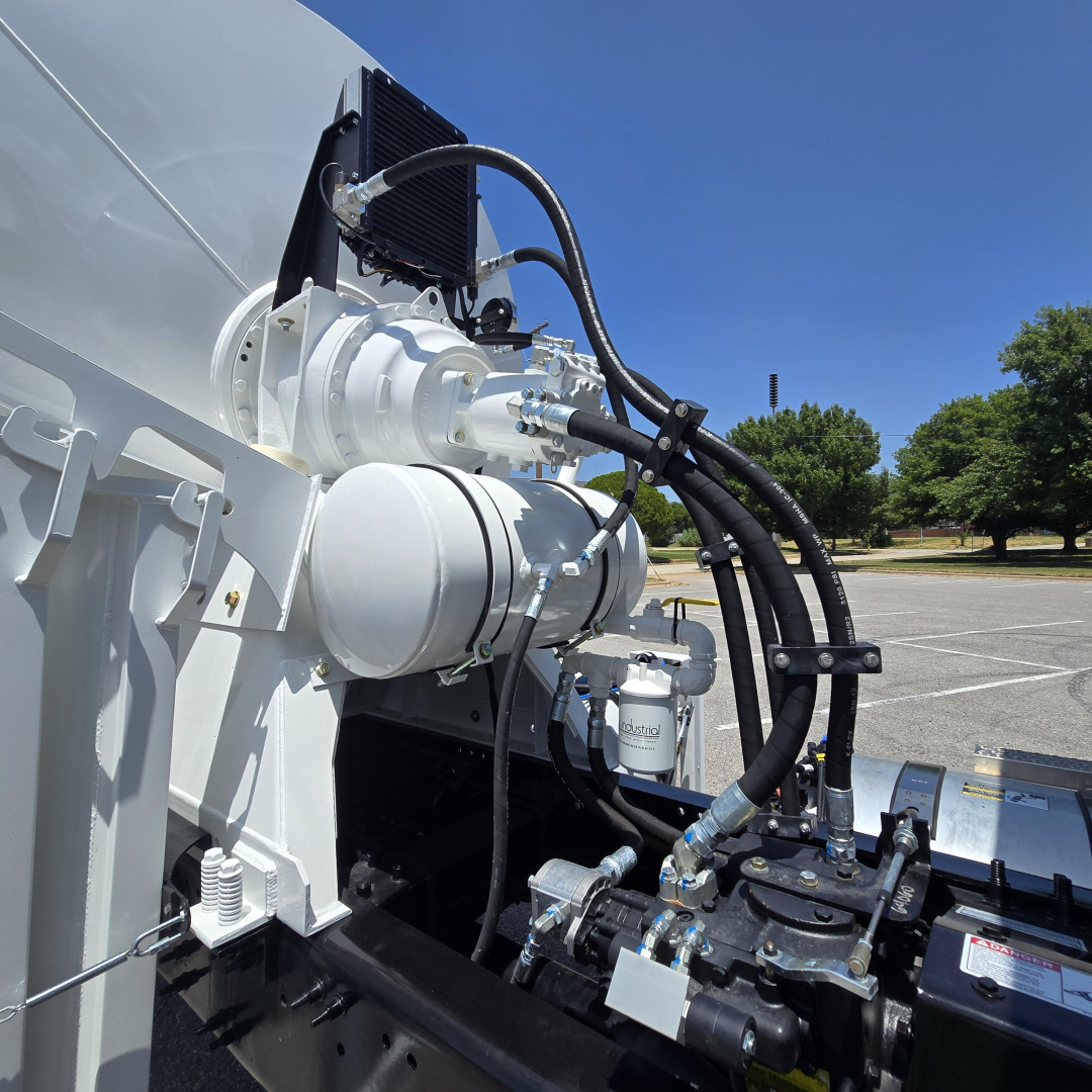 Mixer truck componentry with hydraulic hoses and control valves visible on the chassis of a Mack GR64B mixer, white-painted metal parts and black hoses.