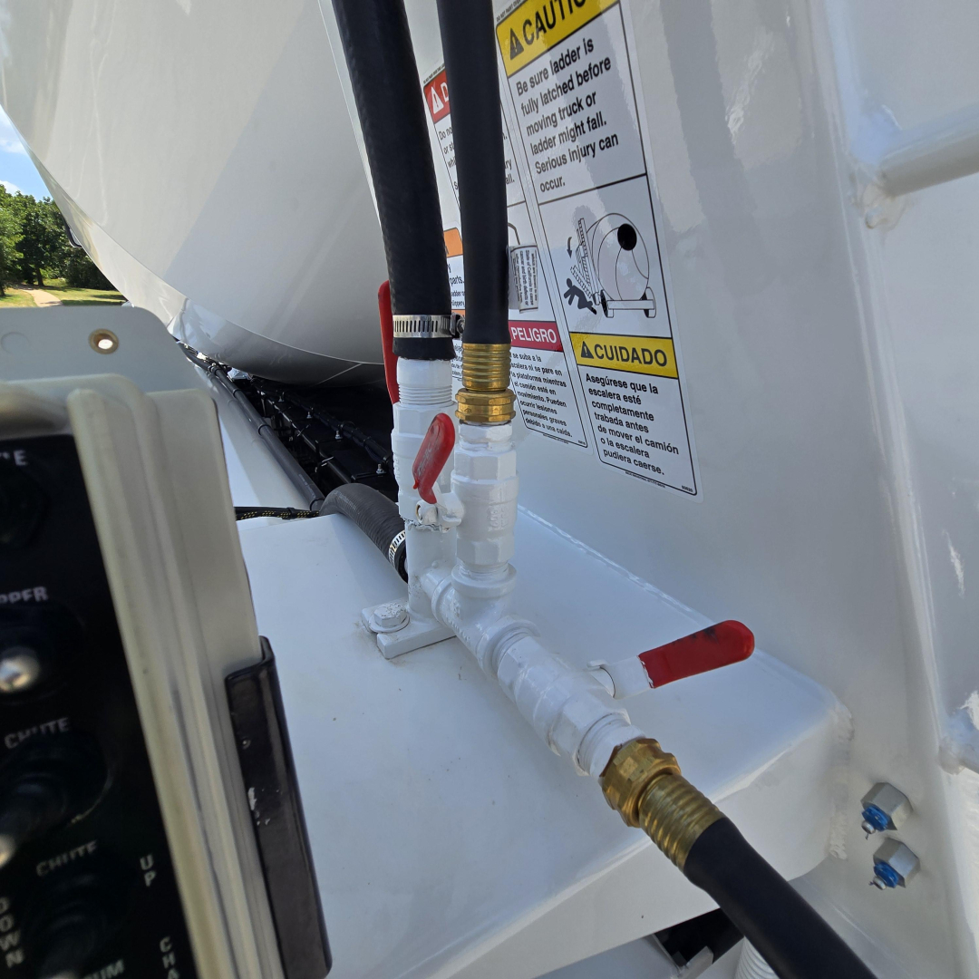Vehicle component close-up showing hydraulic hose fittings and valve controls on a white Mack mixer truck, with safety stickers in the background.