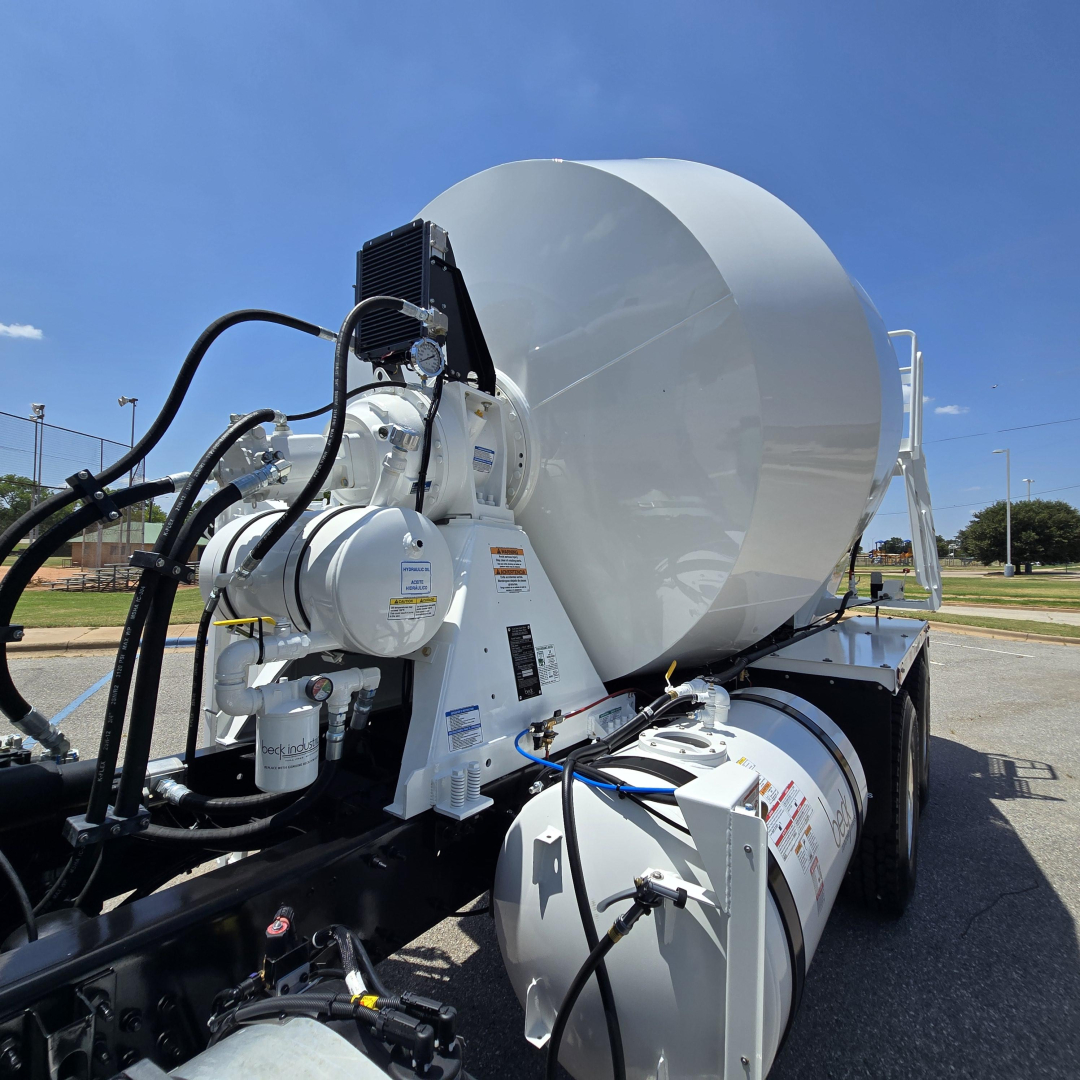 Mixer truck chassis with large white cement mixer drum and hydraulic components, hoses, and control units visible on a sunny day.