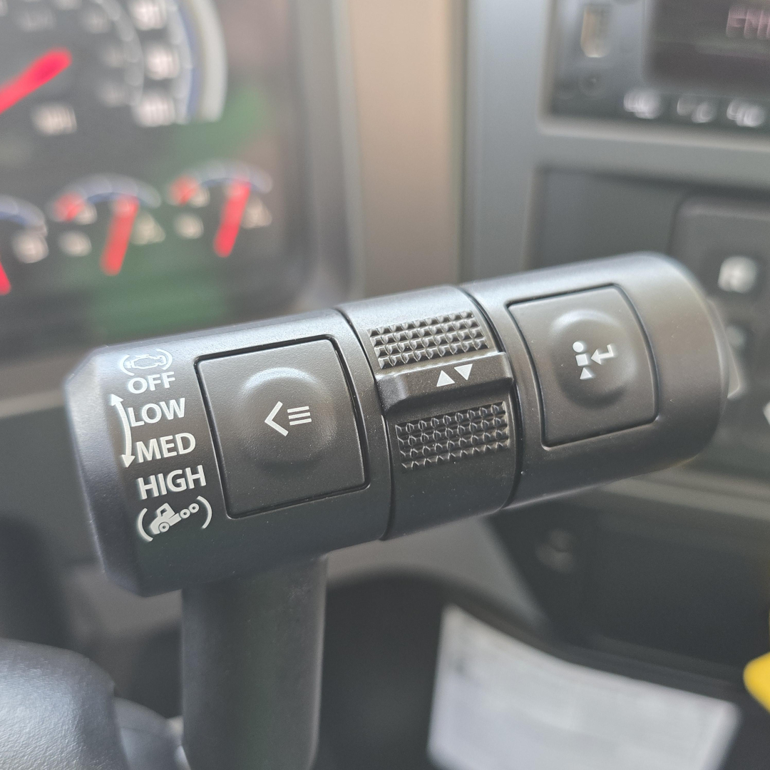 Steering column stalks with multifunction controls in a Mack GR64B truck cabin showing cruise/indicator buttons and a textured grip area.