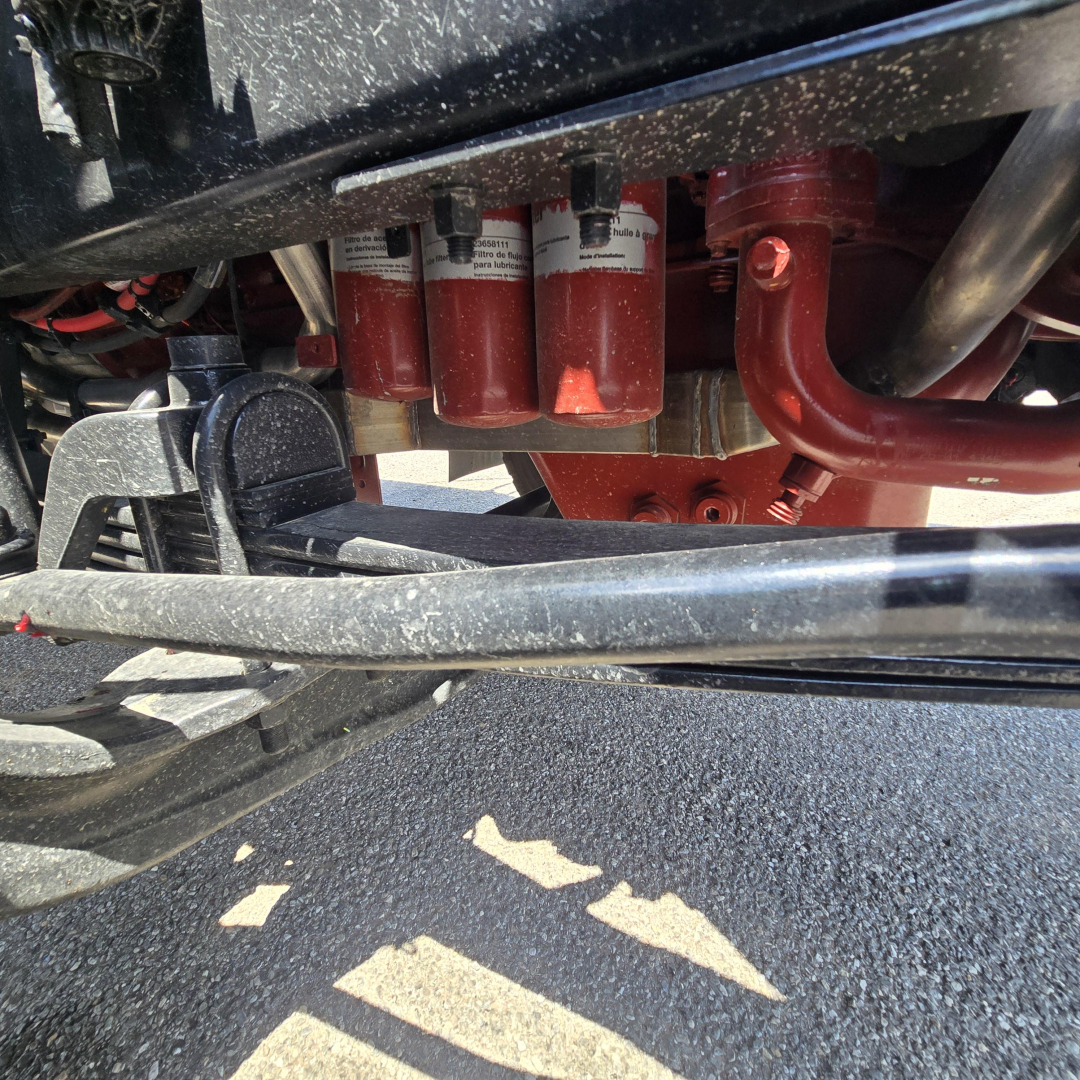 Underbody of a Mack mixer truck showing red coolant/filters and hydraulic lines with black frame and red pipes.