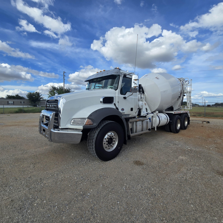 Mack GR64B mixer truck in a gravel lot, white cab with chrome grille, large front bumper, cement mixer drum on back, dual rear axles.
