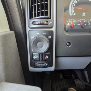 Dashboard area of a Mack truck showing headlight and instrument panel controls, including a round dial, hazard button, and small switch cluster.