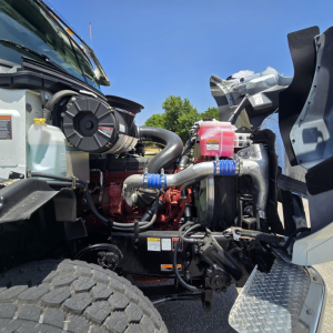 Mack GR64B engine bay showing coolant reservoir, pink reservoir, blue hose clamps, and engine components.