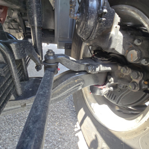 Truck front suspension and steering linkage visible, including a control arm, tie rod, and driveshaft area with engine accessory visible in background.