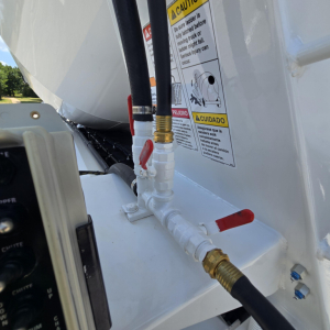 Vehicle component close-up showing hydraulic hose fittings and valve controls on a white Mack mixer truck, with safety stickers in the background.