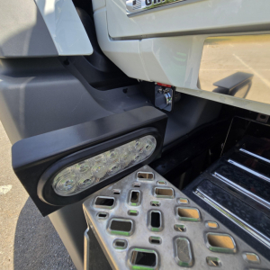 Front left lower cab area of a Mack GR64B mixer showing LED work light, metallic step plate, and partial bumper with VIN plate area.
