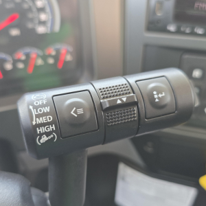 Steering column stalks with multifunction controls in a Mack GR64B truck cabin showing cruise/indicator buttons and a textured grip area.