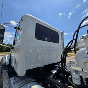 Truck cab area of a white Mack GR64B mixer truck showing driver door window and hydraulic hose assembly near the engine bay.