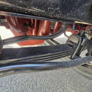 Underbody view of a Mack GR64B mixer truck showing suspension leaf springs and hydraulic lines with red frame components.