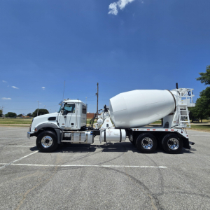 Concrete mixer truck in side view showing cement mixer drum and cab, white color, Mack GR64B chassis with rear wheels and access ladder.