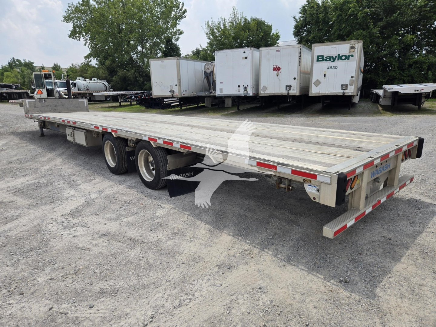 Trailer showing a long flatbed drop deck with three axles and dual tires; background includes several white refrigerated trailers and trees. Visible text includes 'Baylor' on one of the trailers and reflective safety markings on the sides.
