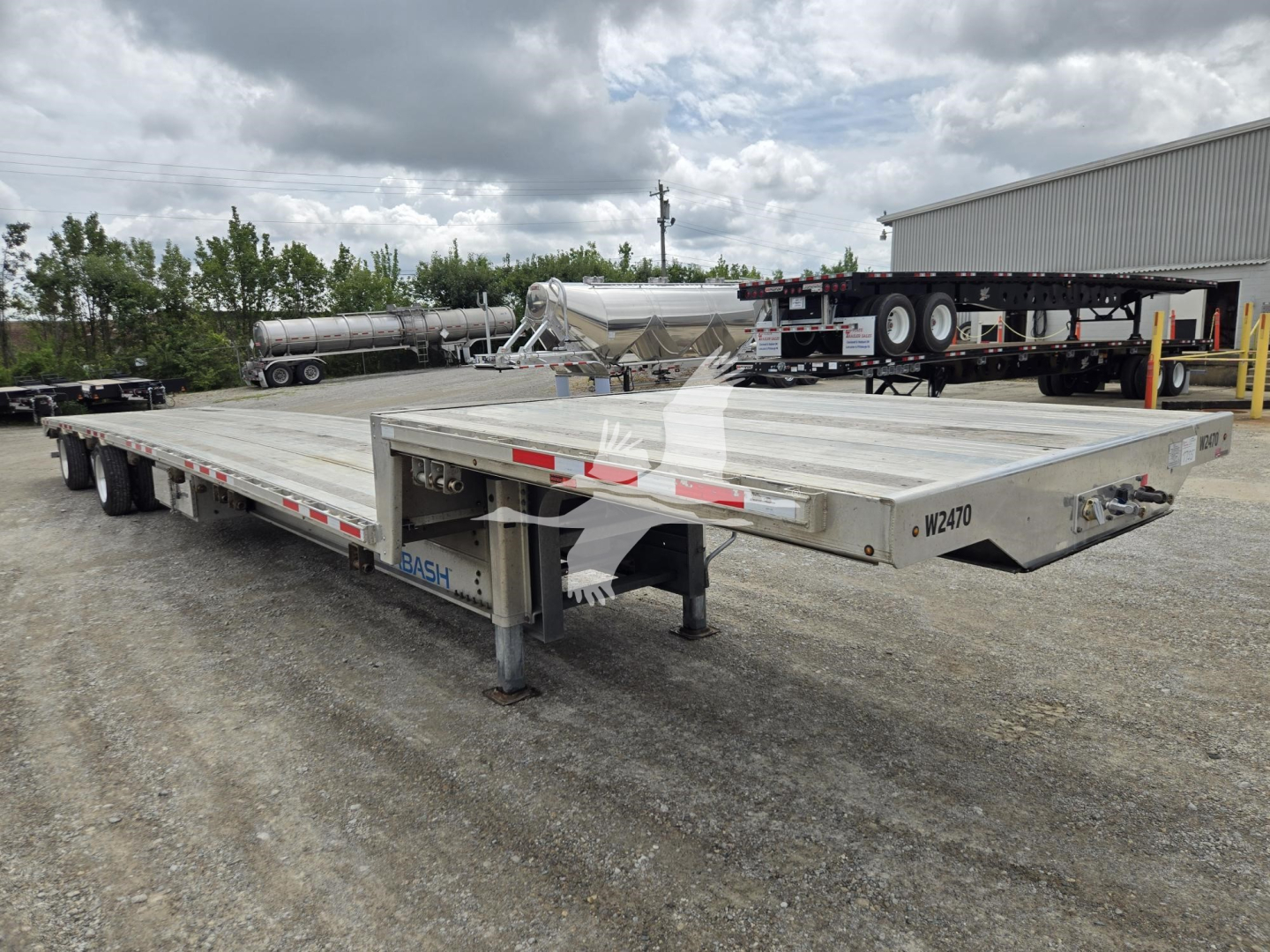 Trailer: aluminum drop deck trailer showing long flat deck and rear ramps, side rails, and landing gear in a gravel lot.
