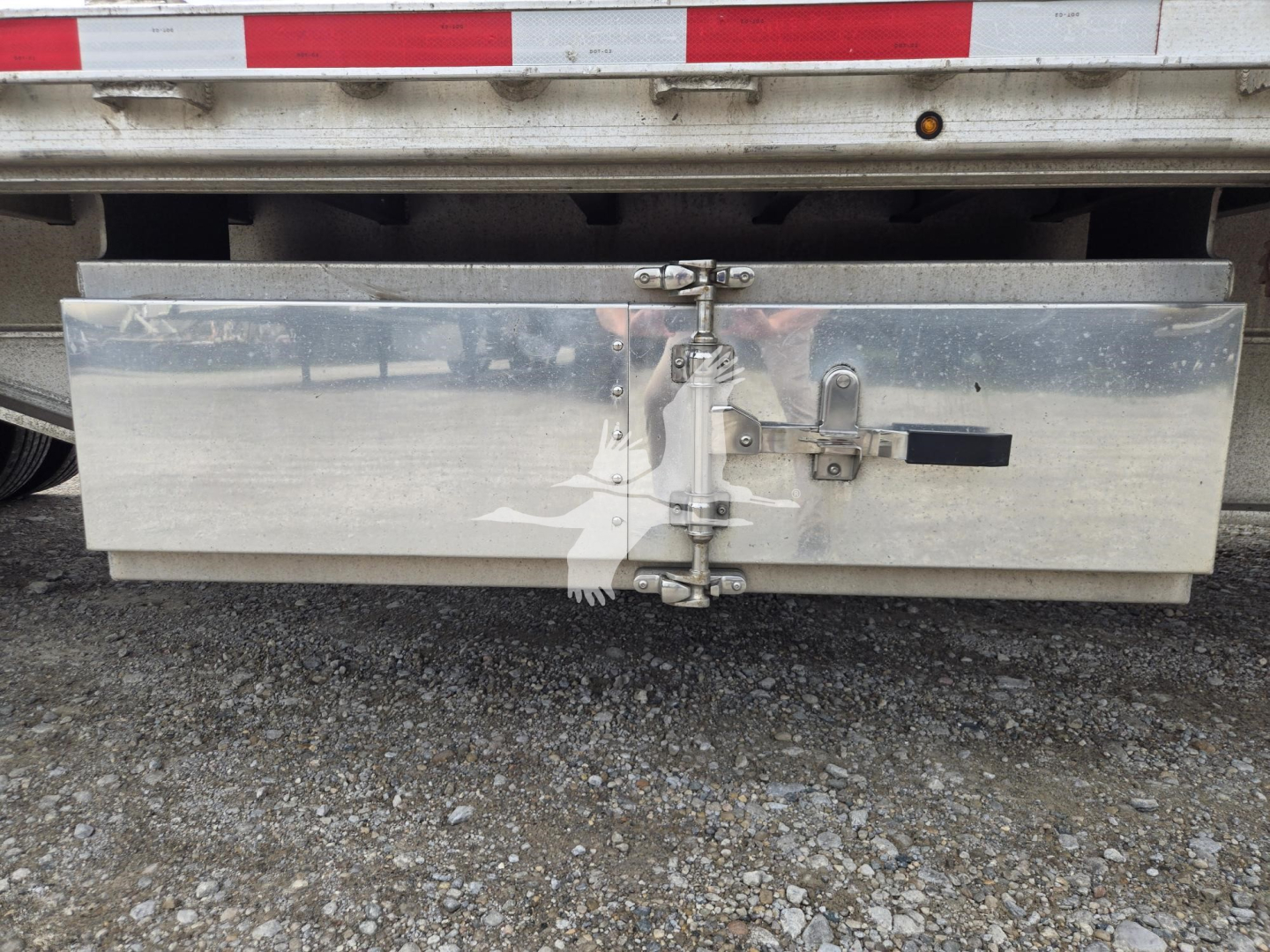 Aluminum drop deck trailer rear showing toolbox door with latch hinges and latch mechanism, silver metal surface, gravel ground.