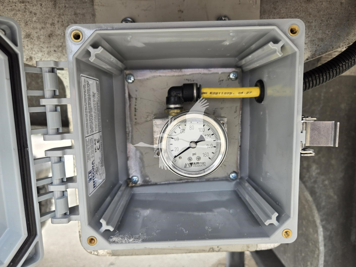 Dial gauge and yellow-handled valve inside a square gray weatherproof box mounted on trailer; visible label and wiring harness; metal hinge and screws.