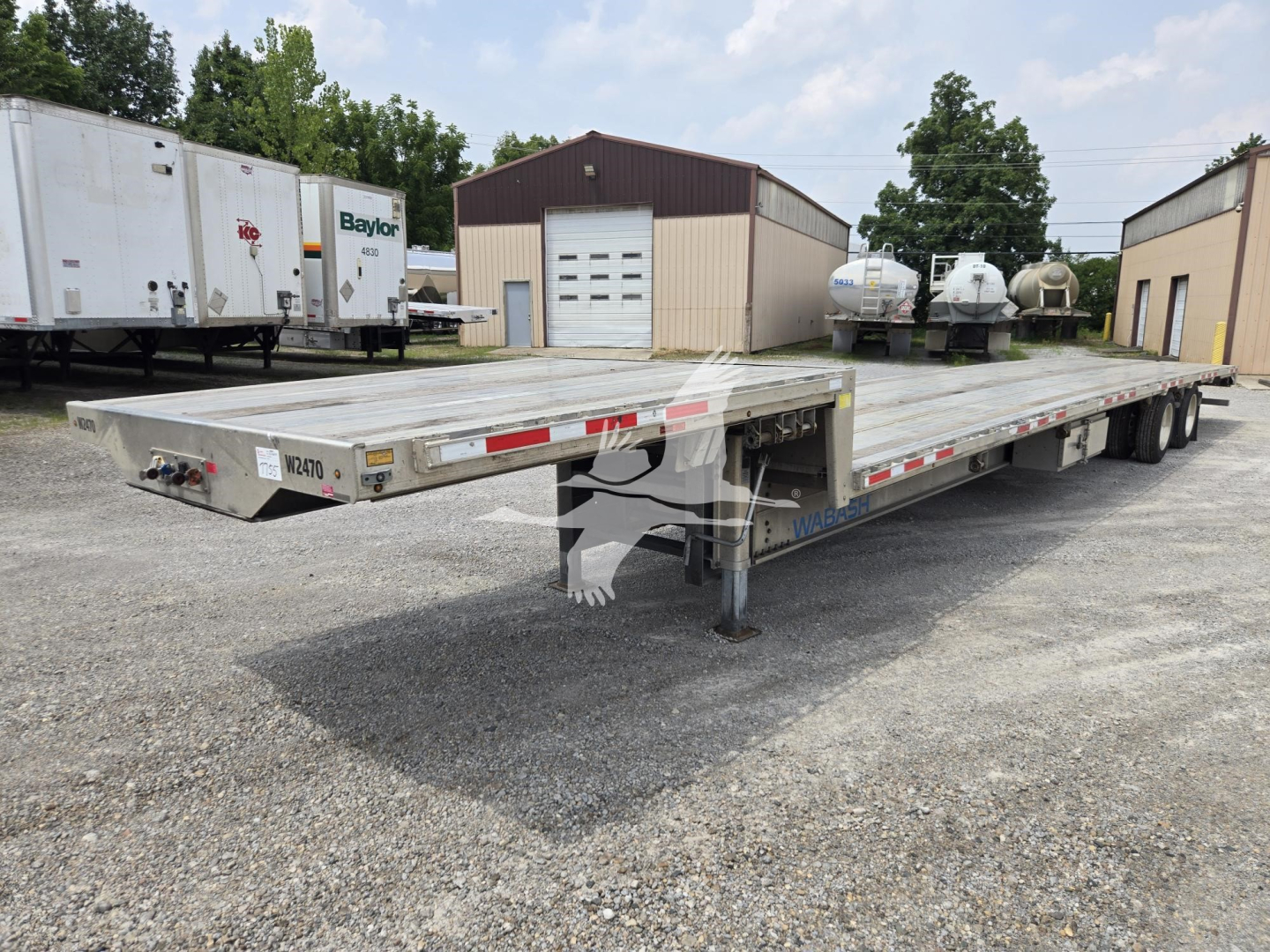 Aluminum drop deck trailer in a gravel lot, viewed from front-left, showing landing gear and deck with reflective tape;