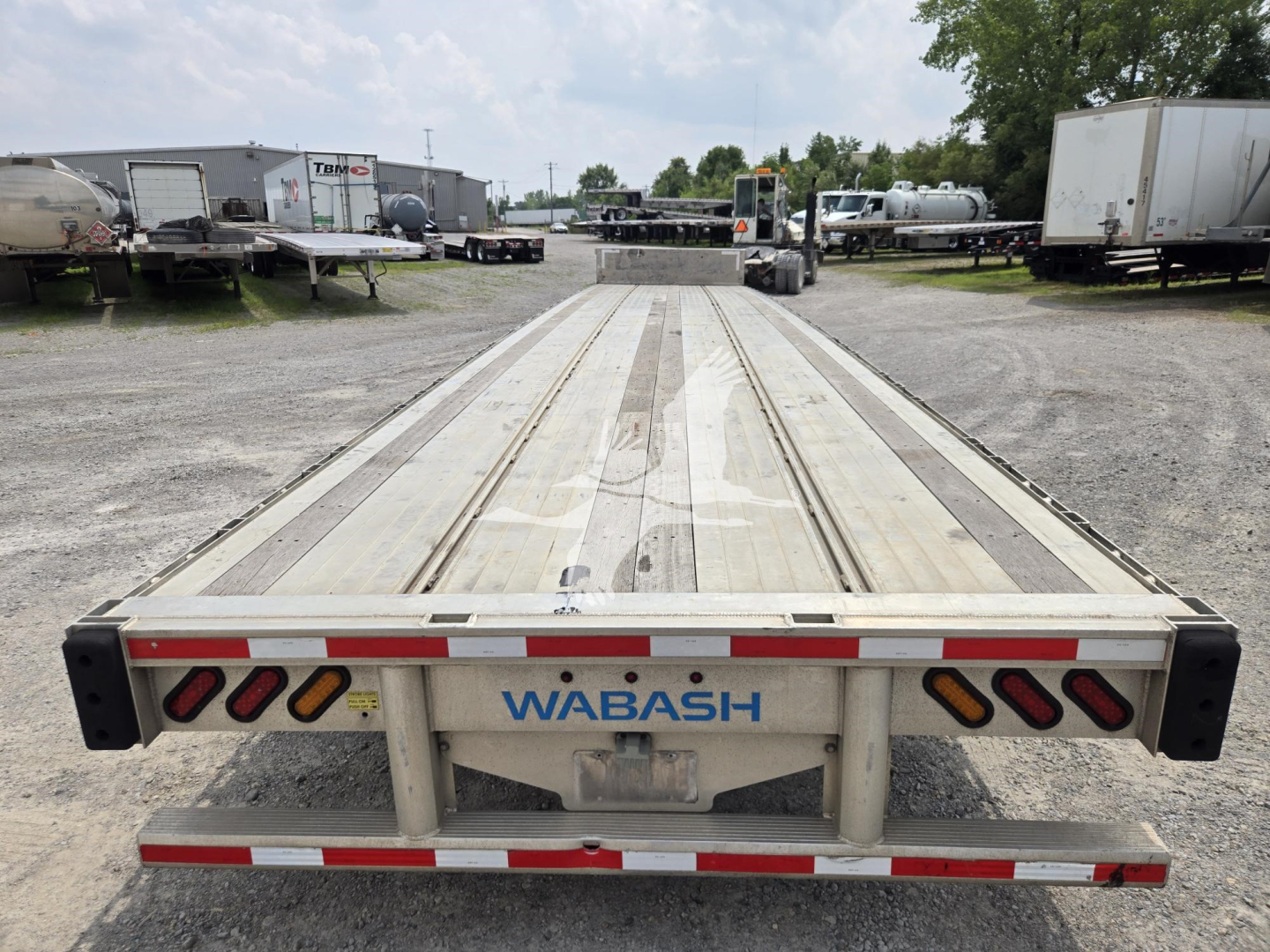 Wabash drop deck trailer viewed from rear, aluminum deck with reflective red/white tape, WABASH branding on center frame, parking lot with trailers in background.