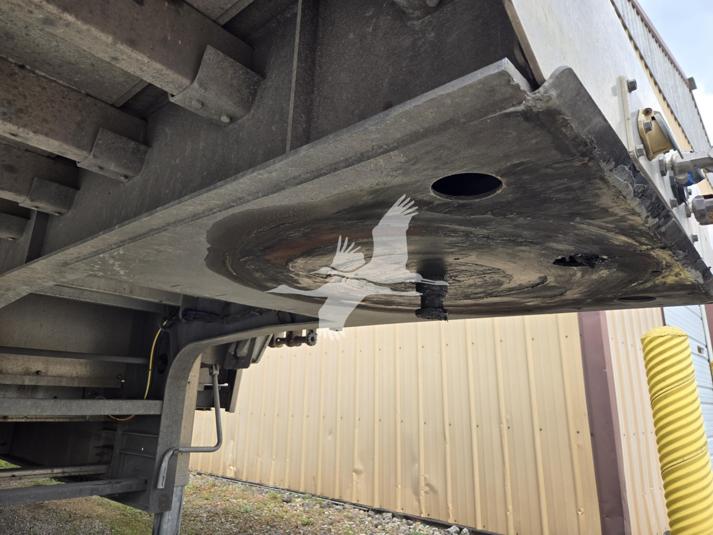 Trailer underside view showing a circular inspection/access hole on the drop deck plate and structural framing with a hydraulic line and edge of platform.