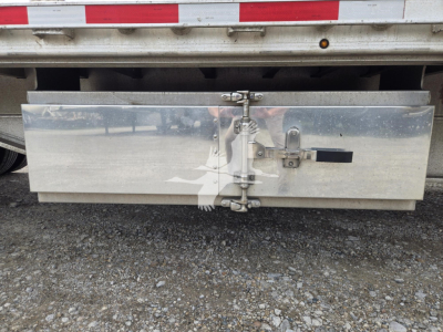 Aluminum drop deck trailer rear showing toolbox door with latch hinges and latch mechanism, silver metal surface, gravel ground.