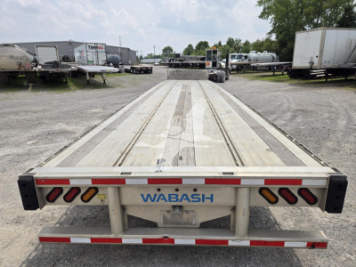 Wabash drop deck trailer viewed from rear, aluminum deck with reflective red/white tape, WABASH branding on center frame, parking lot with trailers in background.