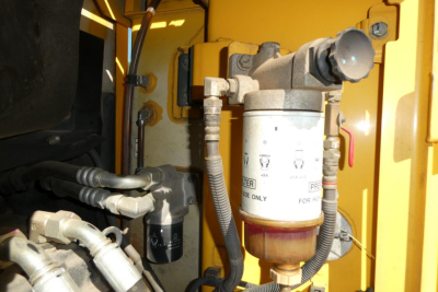 Komatsu PC128US-8 excavator hydraulic filter assembly and adjacent hoses in yellow machine. Visible filter housing with labels and fittings, hydraulic lines and connectors.