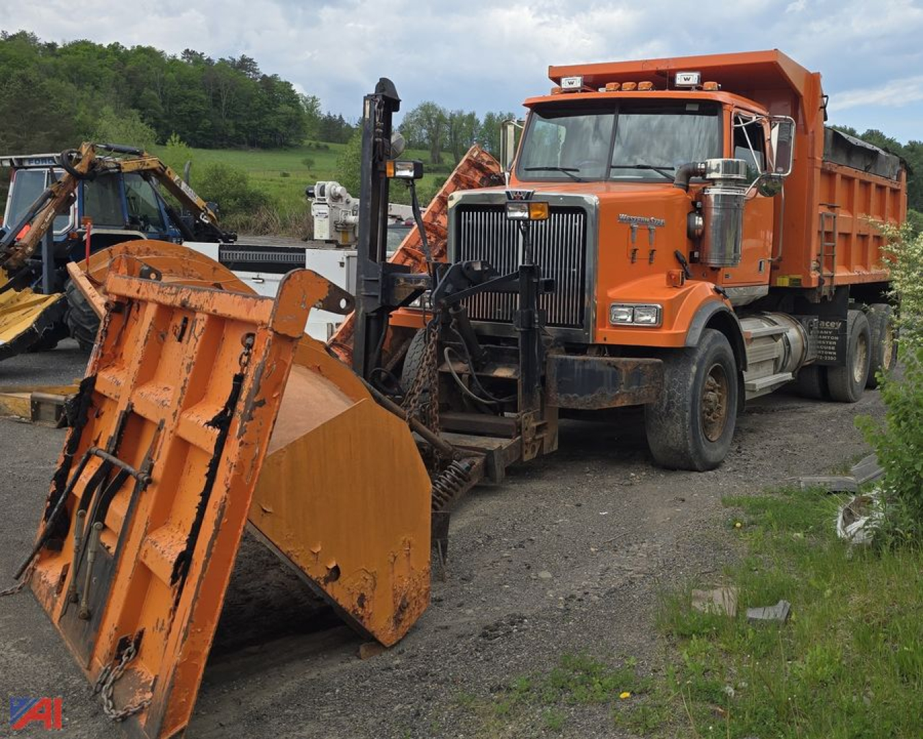 Dump truck with plow attachment in a field, orange Western Star 4900 SF with large front bumper; snow/ice plow blade in foreground and dirt/gravel surface.