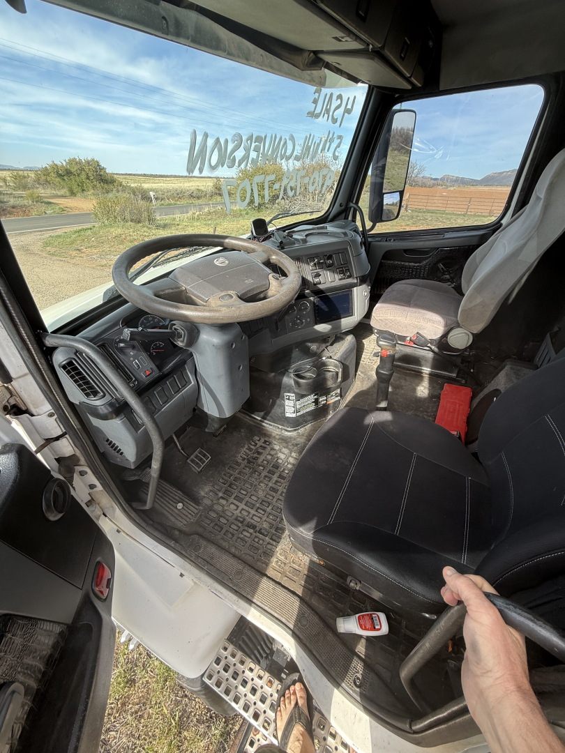 Cab interior of a Volvo truck showing steering wheel, dashboard, pedals, and driver's seat with a hand on a grip; window with writing on glass and desert landscape outside.