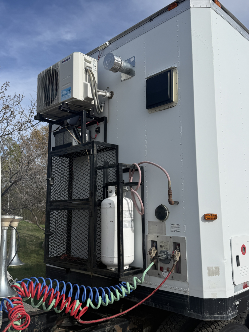 Truck utility area with external A/C condenser, white refrigerator unit, hoses, and control panel on a Volvo VNL trailer unit; visible pipes, wires, and a small window.