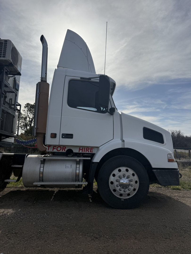 Truck cab side view of a 2005 Volvo VNL64T300 with white exterior, tall sleeper, large exhaust stack, and front wheel visible.